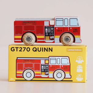 A red and white wooden toy fire truck with light blue windows sits on top of its yellow retail box. The box features the text 'GT270 QUINN' and 'Limited Edition' along with an illustration of the fire truck.