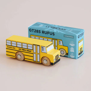 A yellow wooden school bus toy by Le Toy Van placed in front of its blue packaging box on a light beige background.