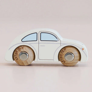 A small wooden toy car shaped like a Volkswagen Beetle, with an off-white painted body and natural wood wheels featuring the word 'Volkswagen' engraved on them, set against a soft pink background.