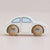 A small wooden toy car shaped like a Volkswagen Beetle, with an off-white painted body and natural wood wheels featuring the word 'Volkswagen' engraved on them, set against a soft pink background.