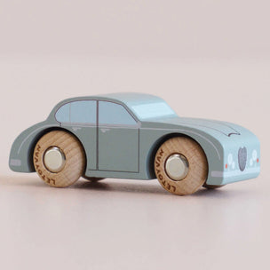 A small wooden toy car in a dusty light blue-green color with natural wood wheels and silver hubcaps, photographed on a plain light beige background.