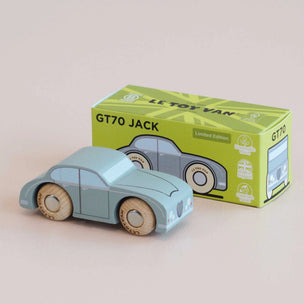 A light blue wooden toy car with natural wood wheels, shown in front of its lime green and yellow packaging box on a neutral beige background.