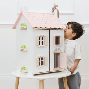 A young boy in a white shirt and gray pants looks up at a large wooden dollhouse table with a pink roof, displayed on a small white table in a bright room.