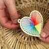 Heart-shaped wooden ornament with colorful string design held by hands on a woven surface