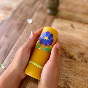 Hand holding a yellow candle object with floral design on a wooden surface with flowers in the background