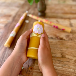 Hand holding a yellow cylindrical container with floral design on a wooden surface with flowers in the background