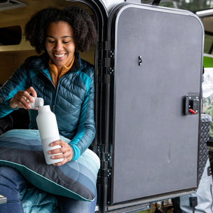 Person sitting in an open vehicle with a water bottle, surrounded by camping gear.