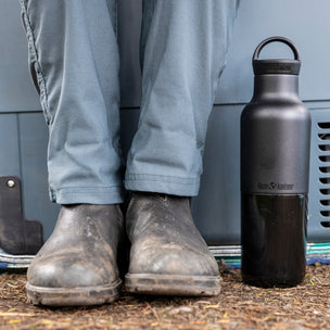 Person wearing gray pants and boots with a black water bottle next to a vehicle.