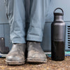 Person wearing gray pants and boots with a black water bottle next to a vehicle.