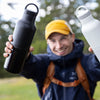 Person holding two water bottles outdoors with a blurred natural background