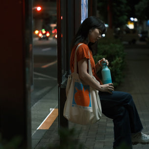 Person sitting on a bench at night holding a water bottle with a city street in the background.