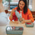 Woman in an orange shirt holding a green water bottle at a table with a phone and container.