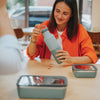 Woman in an orange shirt holding a green water bottle at a table with a phone and container.