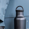 Black water bottle with handle held by a person wearing a blue jacket.