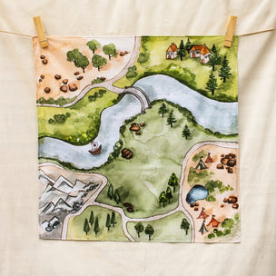 A square fabric tea towel with a watercolor-style landscape illustration, hanging from two wooden clothespins on a string against a cream-colored fabric background.
