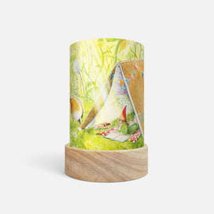 A cylindrical table lamp with a light wood base and a printed fabric shade featuring a whimsical camping scene with a small green gnome, a teepee tent, and a dog, shown on a light gray background.