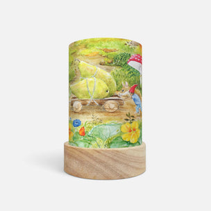 A cylindrical table lamp with a light wood base and a printed fabric shade featuring a whimsical garden scene with a pear cart, flowers, and a bird, shown on a plain light gray background.