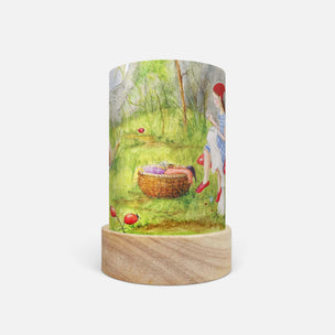 A cylindrical table lamp with a light natural wood base and a printed wrap-around cylinder body featuring a watercolor-style illustration of a girl in a red hat and dress standing in a grassy field near a basket.