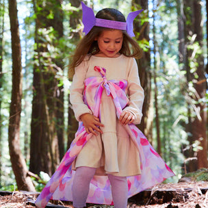 Young girl in a forest wearing a dress with a large pink bow and purple headband.