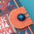 A close-up of a children's book cover featuring a whimsical monster illustration, with an orange bookmark or tab attached by blue elastic bands and a round blue button with a star design.