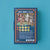 The packaging box of the 'Magic World Puzzle' by the brand 'hapyi' is shown against a solid teal-blue background. The box is dark blue with an orange border and features a colorful, highly detailed illustration of a magical shop scene on the front.