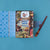 A flat-lay view of a 'Magic World Puzzle' box on a solid blue background, accompanied by a blue star-patterned folder and a wooden wand.
