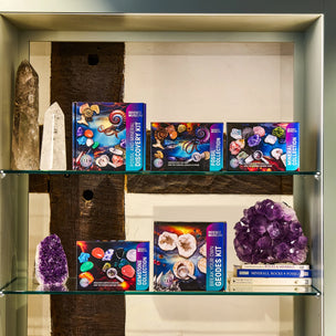 A glass display shelf showcasing crystal and gemstone kits from Discovery Mineral Company, including 'The Great Stone Collection' and 'Geodes Kit', alongside physical crystals like amethyst and a quartz point.