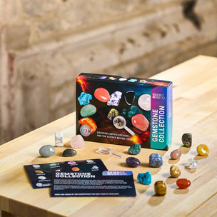 Gemstone collection kit box displayed on a light wood table with several polished and raw gemstones scattered around it. The box reads 'Gemstone Collection' and 'Science Museum,' with a small card in front showing 'Gemstone ID.