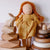 A soft fabric doll with orange-brown braided hair and a mustard yellow dress, posed among natural wooden toys including a rabbit block, geometric blocks, and a ring stack against a light wall.
