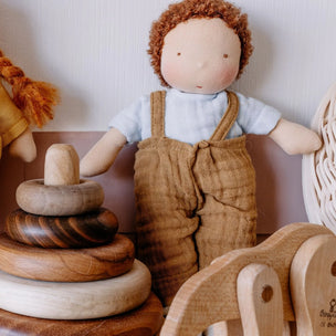 A soft Waldorf fabric doll with curly brown hair, a light blue shirt, and tan overalls sits among wooden toys, including a stack of round pegs and a wooden horse.