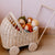 A natural wicker baby carriage toy with wooden wheels and a push handle, filled with soft fabric dolls, set against a light pink paneled wall and wooden floor.