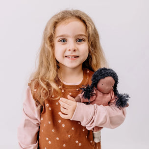 A young girl with long blonde hair smiles while holding a handmade doll with dark curly hair. She is wearing a brown patterned pinafore-style top over a light pink long-sleeve shirt, set against a plain light gray studio background.
