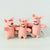 Three pink felt pig toys standing in front of a small wooden box with white tags, photographed against a clean light gray studio background.
