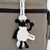 A handmade Shaun the Sheep plush keychain charm with a black and white wool-like body, attached to a beige canvas bag with a black zipper. The keychain includes a white hanging ribbon printed with 'Shaun the Sheep' and 'the Sheep'.