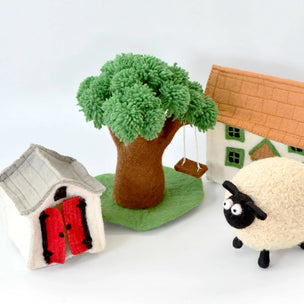 A set of felt playhouse accessories including a green tree with a swing, a white house with a red door, a white house with an orange roof, and a cream-colored sheep with a black face, arranged on a white background.