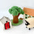 A set of felt playhouse accessories including a green tree with a swing, a white house with a red door, a white house with an orange roof, and a cream-colored sheep with a black face, arranged on a white background.