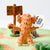 A handmade felt toy of a fluffy orange sheep with white hooves, holding a sign that reads 'Shaun the Sheep'. It is displayed on a green and orange felt path with a wooden sign reading 'Welcome to Messy Bottom Farm' in a miniature outdoor setting.