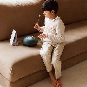 Child playing with a small drum on a beige couch in a bright room.