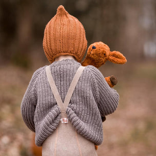 Child wearing a knitted outfit with a hood and suspenders, holding a toy, in a natural setting.