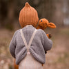 Child wearing a knitted outfit with a hood and suspenders, holding a toy, in a natural setting.