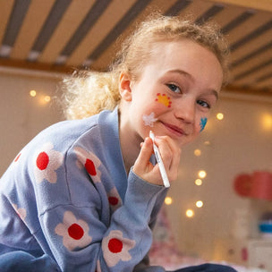 Young girl with face paint holding a marker, sitting at a table with a colorful background.