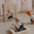 A close-up of a wooden toy figure of an older man sitting in a wooden chair next to a small doll, set on a beige carpet in a softly blurred children's play area.