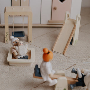 A set of wooden children's playground toys arranged on a beige textured floor, including a swing set, a slide, a seesaw, and small character figures.