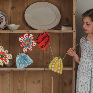Child holding a colorful paper garland in front of a wooden shelf with plates.