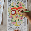 Coloring page of a floral skull with 'Happy Halloween' text, being colored by a hand.