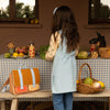Woman standing in a rustic outdoor setting with a table full of fruits and vegetables, wearing a light blue apron.