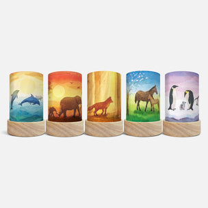 Five cylindrical decorative lamps with light wood bases, each featuring a different watercolor animal scene: dolphins, an elephant and calf, a fox, a horse and foal, and penguins, shown on a light gray background.
