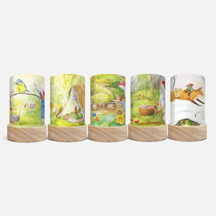 Five cylindrical decorative items with light wood bases, each featuring a different whimsical illustrated scene in soft pastel colors, arranged side by side on a light gray background.