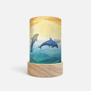 A cylindrical table lamp with a light wood base and a printed wrap featuring two dolphins swimming over blue water under a yellow sun, shown on a light gray background.