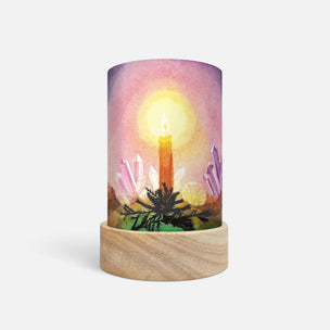 A cylindrical table lamp with a printed watercolor-style design of a glowing candle among crystals, set on a light natural wood base against a plain white background.